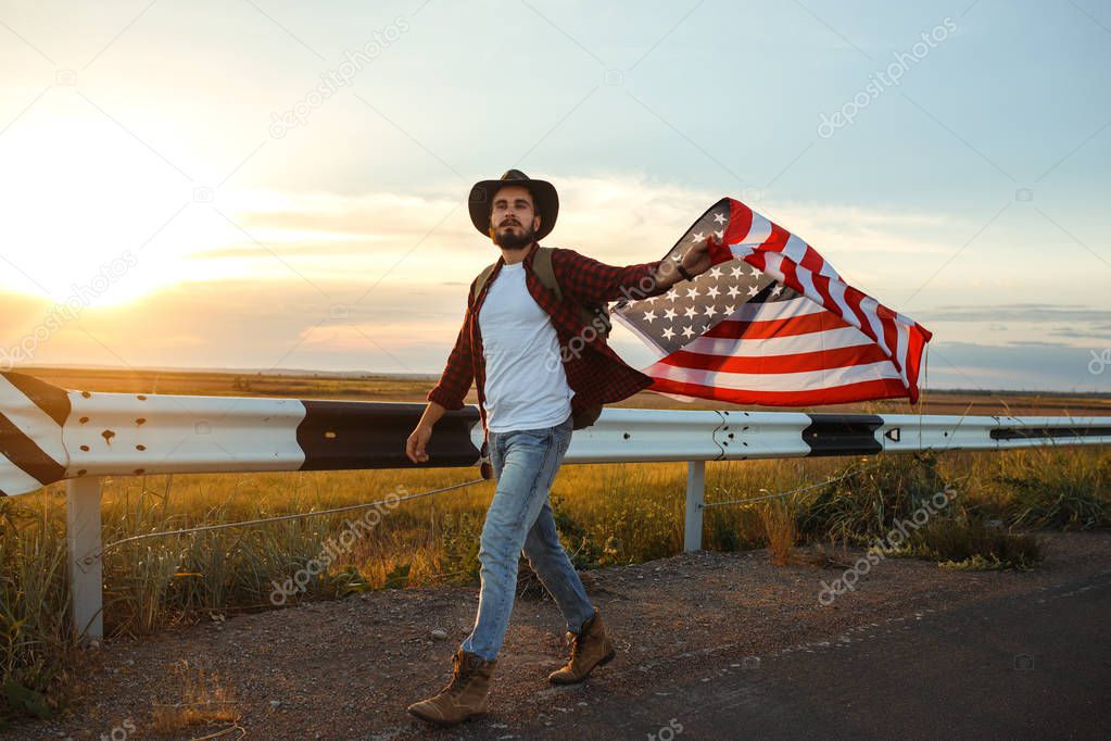 4th of July. Fourth of July. American with the national flag. American Flag. Independence Day. Patriotic holiday. The man is wearing a hat, a backpack, a shirt and jeans. Beautiful sunset light. 