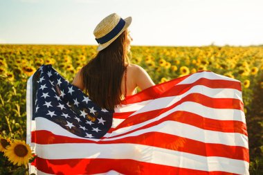 Ayçiçeği tarlasında Amerikan bayrağı olan güzel bir kız. 4 Temmuz. 4 Temmuz. Özgürlük. Gün batımı ışığı Kız gülümsüyor. Güzel gün batımı. Bağımsızlık Günü. Vatansever tatil.