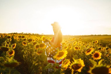 Ayçiçeği tarlasında Amerikan bayrağı olan güzel bir kız. 4 Temmuz. 4 Temmuz. Özgürlük. Gün batımı ışığı Kız gülümsüyor. Güzel gün batımı. Bağımsızlık Günü. Vatansever tatil.