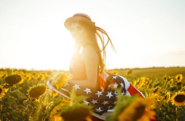 Ayçiçeği tarlasında Amerikan bayrağı olan güzel bir kız. 4 Temmuz. 4 Temmuz. Özgürlük. Gün batımı ışığı Kız gülümsüyor. Güzel gün batımı. Bağımsızlık Günü. Vatansever tatil.