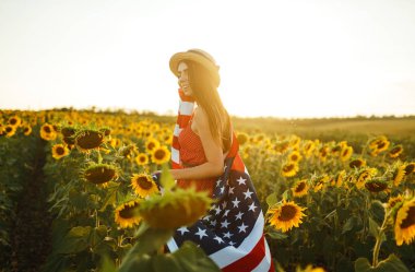 Ayçiçeği tarlasında Amerikan bayrağı olan güzel bir kız. 4 Temmuz. 4 Temmuz. Özgürlük. Gün batımı ışığı Kız gülümsüyor. Güzel gün batımı. Bağımsızlık Günü. Vatansever tatil.
