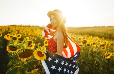 Ayçiçeği tarlasında Amerikan bayrağı olan güzel bir kız. 4 Temmuz. 4 Temmuz. Özgürlük. Gün batımı ışığı Kız gülümsüyor. Güzel gün batımı. Bağımsızlık Günü. Vatansever tatil.