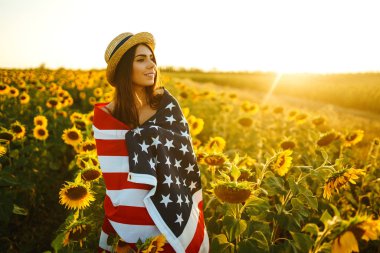 Ayçiçeği tarlasında Amerikan bayrağı olan güzel bir kız. 4 Temmuz. 4 Temmuz. Özgürlük. Gün batımı ışığı Kız gülümsüyor. Güzel gün batımı. Bağımsızlık Günü. Vatansever tatil.