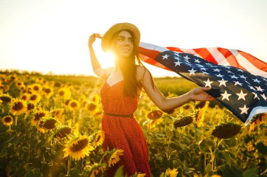Ayçiçeği tarlasında Amerikan bayrağı olan güzel bir kız. 4 Temmuz. 4 Temmuz. Özgürlük. Gün batımı ışığı Kız gülümsüyor. Güzel gün batımı. Bağımsızlık Günü. Vatansever tatil.