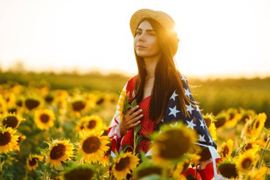 Ayçiçeği tarlasında Amerikan bayrağı olan güzel bir kız. 4 Temmuz. 4 Temmuz. Özgürlük. Gün batımı ışığı Kız gülümsüyor. Güzel gün batımı. Bağımsızlık Günü. Vatansever tatil.