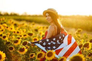 Ayçiçeği tarlasında Amerikan bayrağı olan güzel bir kız. 4 Temmuz. 4 Temmuz. Özgürlük. Gün batımı ışığı Kız gülümsüyor. Güzel gün batımı. Bağımsızlık Günü. Vatansever tatil.