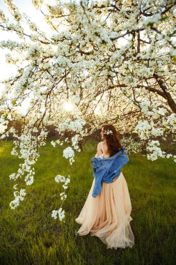 Güzel bir gelinin portresi. Düğün konsepti. Şık bir düğün. Harika gün batımı ışığı. Mavi kot ceket giyiyor, elbise. Bahar çiçekli bahçe..