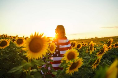 Ayçiçeği tarlasında Amerikan bayrağı olan güzel bir kız. 4 Temmuz. 4 Temmuz. Özgürlük. Gün batımı ışığı Kız gülümsüyor. Güzel gün batımı. Bağımsızlık Günü. Vatansever tatil. 