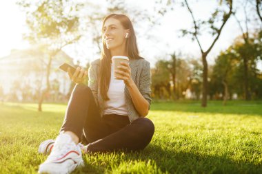 El ve kahve bir telefon ile yeşil çim üzerinde oturan güzel şık kadın görüntü. O kablosuz kulaklıklar üzerinden telefonda konuşuyor. Gün batımı ışığı. Yaşam tarzı konsepti.