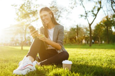 El ve kahve bir telefon ile yeşil çim üzerinde oturan güzel şık kadın görüntü. O kablosuz kulaklıklar üzerinden telefonda konuşuyor. Gün batımı ışığı. Yaşam tarzı konsepti.