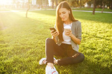 El ve kahve bir telefon ile yeşil çim üzerinde oturan güzel şık kadın görüntü. O kablosuz kulaklıklar üzerinden telefonda konuşuyor. Gün batımı ışığı. Yaşam tarzı konsepti.
