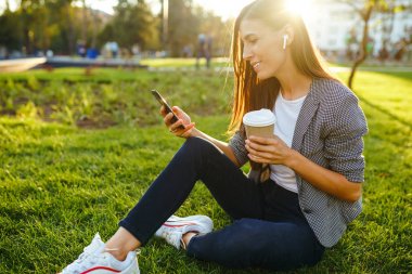 El ve kahve bir telefon ile yeşil çim üzerinde oturan güzel şık kadın görüntü. O kablosuz kulaklıklar üzerinden telefonda konuşuyor. Gün batımı ışığı. Yaşam tarzı konsepti.