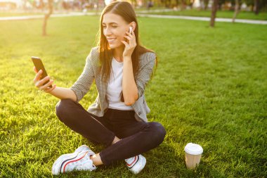 El ve kahve bir telefon ile yeşil çim üzerinde oturan güzel şık kadın görüntü. O kablosuz kulaklıklar üzerinden telefonda konuşuyor. Gün batımı ışığı. Yaşam tarzı konsepti.