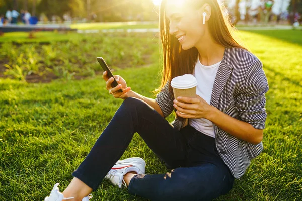 Image of beautiful stylish woman sitting on green grass with a phone in hand and coffee. She is talking on the phone through wireless headphones. Sunset light. Lifestyle concept.