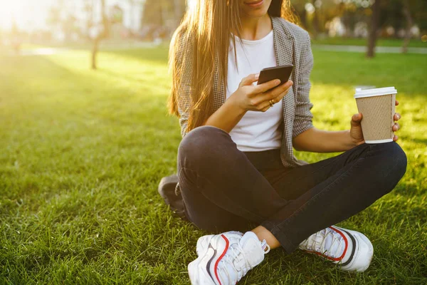 The girl holds the phone and coffee in hand. Image of beautiful stylish woman sitting on green grass with a phone in hand and coffee.  Sunset.  Summertime.