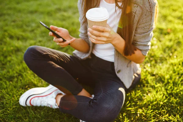 The girl holds the phone and coffee in hand. Image of beautiful stylish woman sitting on green grass with a phone in hand and coffee.  Sunset.  Summertime.