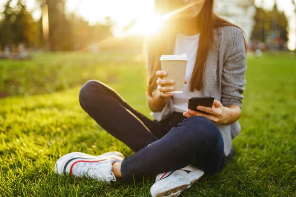 The girl holds the phone and coffee in hand. Image of beautiful stylish woman sitting on green grass with a phone in hand and coffee.  Sunset.  Summertime.