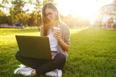 El dizüstü ve kahve ile yeşil çim üzerinde oturan güzel şık kadın görüntü. O kablosuz kulaklıklar üzerinden telefonda konuşuyor. Gün batımı ışığı. Yaşam tarzı konsepti