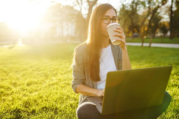 El dizüstü ve kahve ile yeşil çim üzerinde oturan güzel şık kadın görüntü. O kablosuz kulaklıklar üzerinden telefonda konuşuyor. Gün batımı ışığı. Yaşam tarzı konsepti