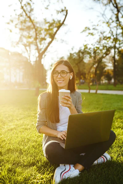 El dizüstü ve kahve ile yeşil çim üzerinde oturan güzel şık kadın görüntü. O kablosuz kulaklıklar üzerinden telefonda konuşuyor. Gün batımı ışığı. Yaşam tarzı konsepti