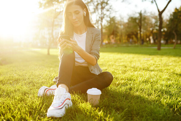 Image of beautiful stylish woman sitting on green grass with laptop and coffee in the hand. She is talking on the phone through wireless headphones. Sunset light. Lifestyle concept