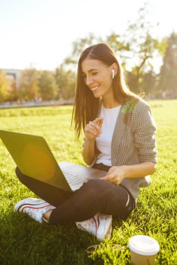 Güzel şık kadın laptop ile yeşil çim üzerinde oturan görüntü o kablosuz kulaklıklar üzerinden telefonda konuşuyor. Gün batımı ışığı. Yaşam tarzı konsepti.