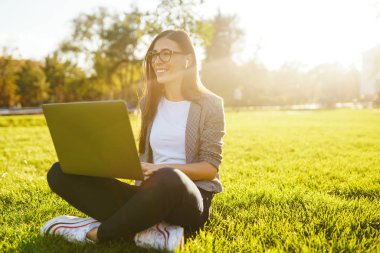 Güzel şık kadın laptop ile yeşil çim üzerinde oturan görüntü o kablosuz kulaklıklar üzerinden telefonda konuşuyor. Gün batımı ışığı. Yaşam tarzı konsepti.