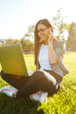 Güzel şık kadın laptop ile yeşil çim üzerinde oturan görüntü o kablosuz kulaklıklar üzerinden telefonda konuşuyor. Gün batımı ışığı. Yaşam tarzı konsepti.