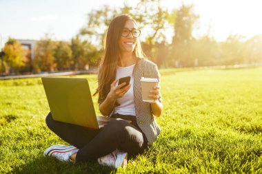 El laptop ve telefon ile yeşil çim üzerinde oturan güzel şık kadın görüntü. O kablosuz kulaklıklar üzerinden telefonda konuşuyor. Gün batımı ışığı. Yaşam tarzı konsepti.