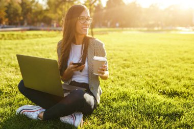 El laptop ve telefon ile yeşil çim üzerinde oturan güzel şık kadın görüntü. O kablosuz kulaklıklar üzerinden telefonda konuşuyor. Gün batımı ışığı. Yaşam tarzı konsepti.