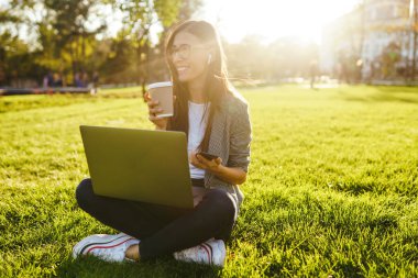 El laptop ve telefon ile yeşil çim üzerinde oturan güzel şık kadın görüntü. O kablosuz kulaklıklar üzerinden telefonda konuşuyor. Gün batımı ışığı. Yaşam tarzı konsepti.