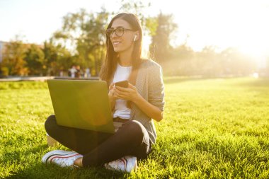 El laptop ve telefon ile yeşil çim üzerinde oturan güzel şık kadın görüntü. O kablosuz kulaklıklar üzerinden telefonda konuşuyor. Gün batımı ışığı. Yaşam tarzı konsepti.