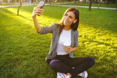 Selfie çekmek için zaman! Elinde telefon ve kahve ile yeşil çim üzerinde oturan güzel şık kadın görüntü. O kablosuz kulaklıklar üzerinden telefonda konuşuyor. Gün batımı ışığı. Yaşam tarzı konsepti