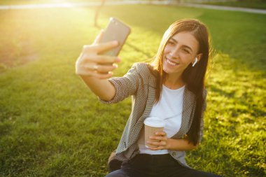Selfie çekmek için zaman! Elinde telefon ve kahve ile yeşil çim üzerinde oturan güzel şık kadın görüntü. O kablosuz kulaklıklar üzerinden telefonda konuşuyor. Gün batımı ışığı. Yaşam tarzı konsepti