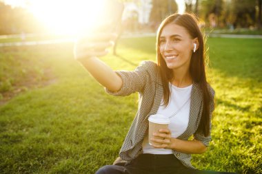 Selfie çekmek için zaman! Elinde telefon ve kahve ile yeşil çim üzerinde oturan güzel şık kadın görüntü. O kablosuz kulaklıklar üzerinden telefonda konuşuyor. Gün batımı ışığı. Yaşam tarzı konsepti