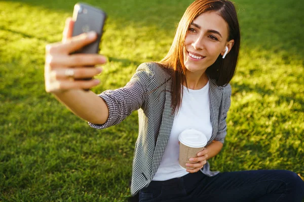 Selfie çekmek için zaman! Elinde telefon ve kahve ile yeşil çim üzerinde oturan güzel şık kadın görüntü. O kablosuz kulaklıklar üzerinden telefonda konuşuyor. Gün batımı ışığı. Yaşam tarzı konsepti
