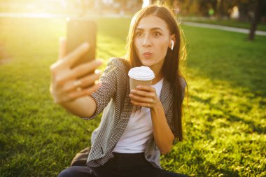 Selfie çekmek için zaman! Elinde telefon ve kahve ile yeşil çim üzerinde oturan güzel şık kadın görüntü. O kablosuz kulaklıklar üzerinden telefonda konuşuyor. Gün batımı ışığı. Yaşam tarzı konsepti