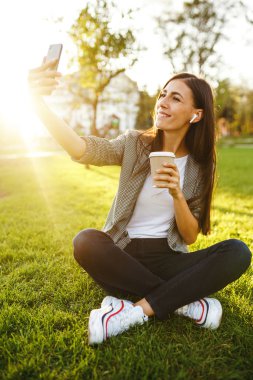 Selfie çekmek için zaman! Elinde telefon ve kahve ile yeşil çim üzerinde oturan güzel şık kadın görüntü. O kablosuz kulaklıklar üzerinden telefonda konuşuyor. Gün batımı ışığı. Yaşam tarzı konsepti