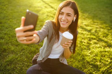 Selfie çekmek için zaman! Elinde telefon ve kahve ile yeşil çim üzerinde oturan güzel şık kadın görüntü. O kablosuz kulaklıklar üzerinden telefonda konuşuyor. Gün batımı ışığı. Yaşam tarzı konsepti