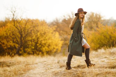 Paltolu güzel, genç, şık bir kız sonbaharda parkta yürüyor. Kız yeşil bir ceket ve kırmızı bir şapka giyiyor. Güzel bir akşam. Sonbahar modası. Yaşam tarzı. Yüksek moda portresi.