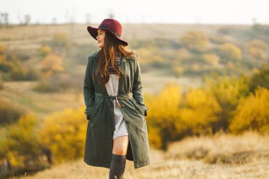 Paltolu güzel, genç, şık bir kız sonbaharda parkta yürüyor. Kız yeşil bir ceket ve kırmızı bir şapka giyiyor. Güzel bir akşam. Sonbahar modası. Yaşam tarzı. Yüksek moda portresi.