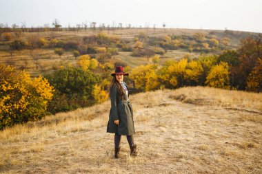 Paltolu güzel, genç, şık bir kız sonbaharda parkta yürüyor. Kız yeşil bir ceket ve kırmızı bir şapka giyiyor. Güzel bir akşam. Sonbahar modası. Yaşam tarzı. Yüksek moda portresi.
