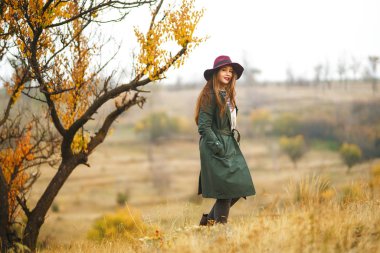 Paltolu güzel, genç, şık bir kız sonbaharda parkta yürüyor. Kız yeşil bir ceket ve kırmızı bir şapka giyiyor. Güzel bir akşam. Sonbahar modası. Yaşam tarzı. Yüksek moda portresi.