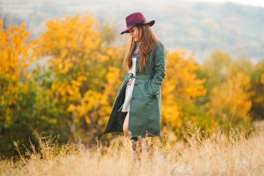 Paltolu güzel, genç, şık bir kız sonbaharda parkta yürüyor. Kız yeşil bir ceket ve kırmızı bir şapka giyiyor. Güzel bir akşam. Sonbahar modası. Yaşam tarzı. Yüksek moda portresi.