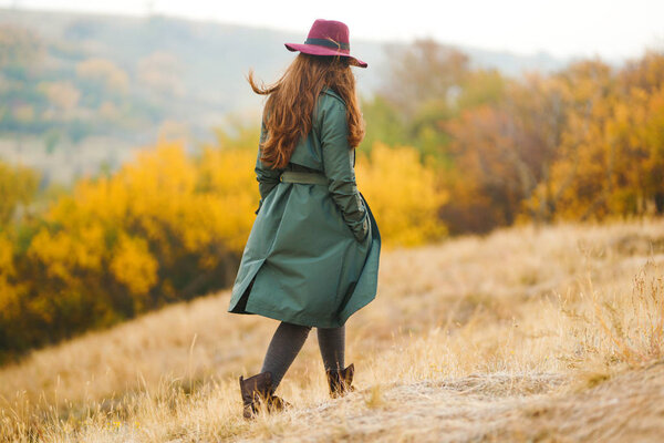 Beautiful young stylish girl in a coat walks in the autumn in the park. The girl is dressed in a green coat and a red hat. Beautiful evening. Autumn fashion. Lifestyle. High fashion portrait.