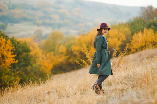 Beautiful young stylish girl in a coat walks in the autumn in the park. The girl is dressed in a green coat and a red hat. Beautiful evening. Autumn fashion. Lifestyle. High fashion portrait.