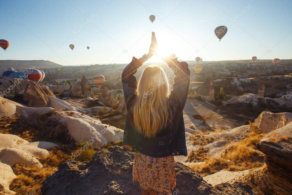 Young attractive girl in a hat stands on the mountain with flying air balloons on the background. Finger pointing girl in the sunrise. View from the back. Famous tourist Turkish region Cappadocia.