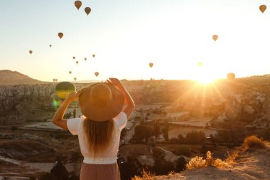 Arka planda uçan balonlarla dağda duran şapkalı güzel genç ve çekici bir kız. Güneşin doğuşundaki kız. Arkadan bak. Ünlü Türk bölgesi Kapadokya.
