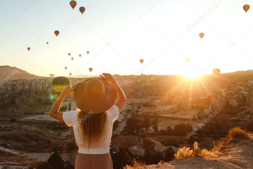 Beautiful young attractive girl in a hat stands on the mountain with flying air balloons on the background. Girl in the sunrise. View from the back. Famous tourist Turkish region Cappadocia.