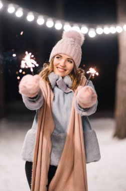 Elinde maytap olan güzel bir kız. Ormanda mutlu kış zamanları. Kız mavi bir ceket giymiş, açık renk bir şapka, atkı ve eldiven takmış. Şenlikli çelenk ışıkları. Noel, yeni yıl.